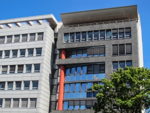Two adjacent office blocks with a mix of white and gray facades, featuring rows of windows and a glimpse of greenery under a bright sky.